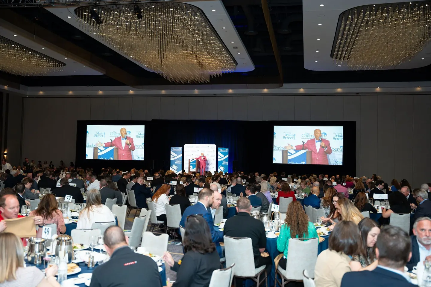 A speaker addresses a conference audience beneath chandeliers